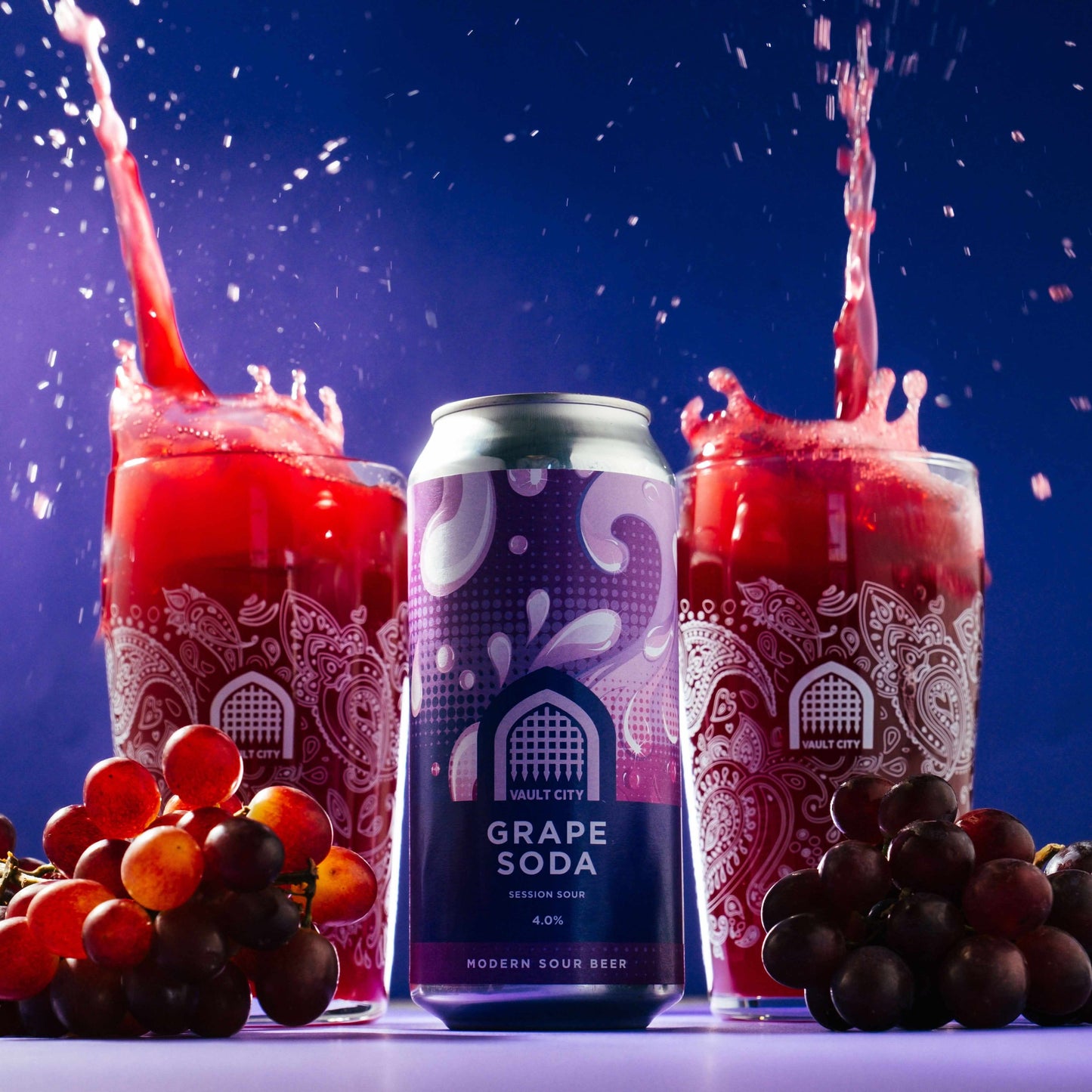 Grape soda session sour beer can surrounded by grapes and two splashing glasses on a table.