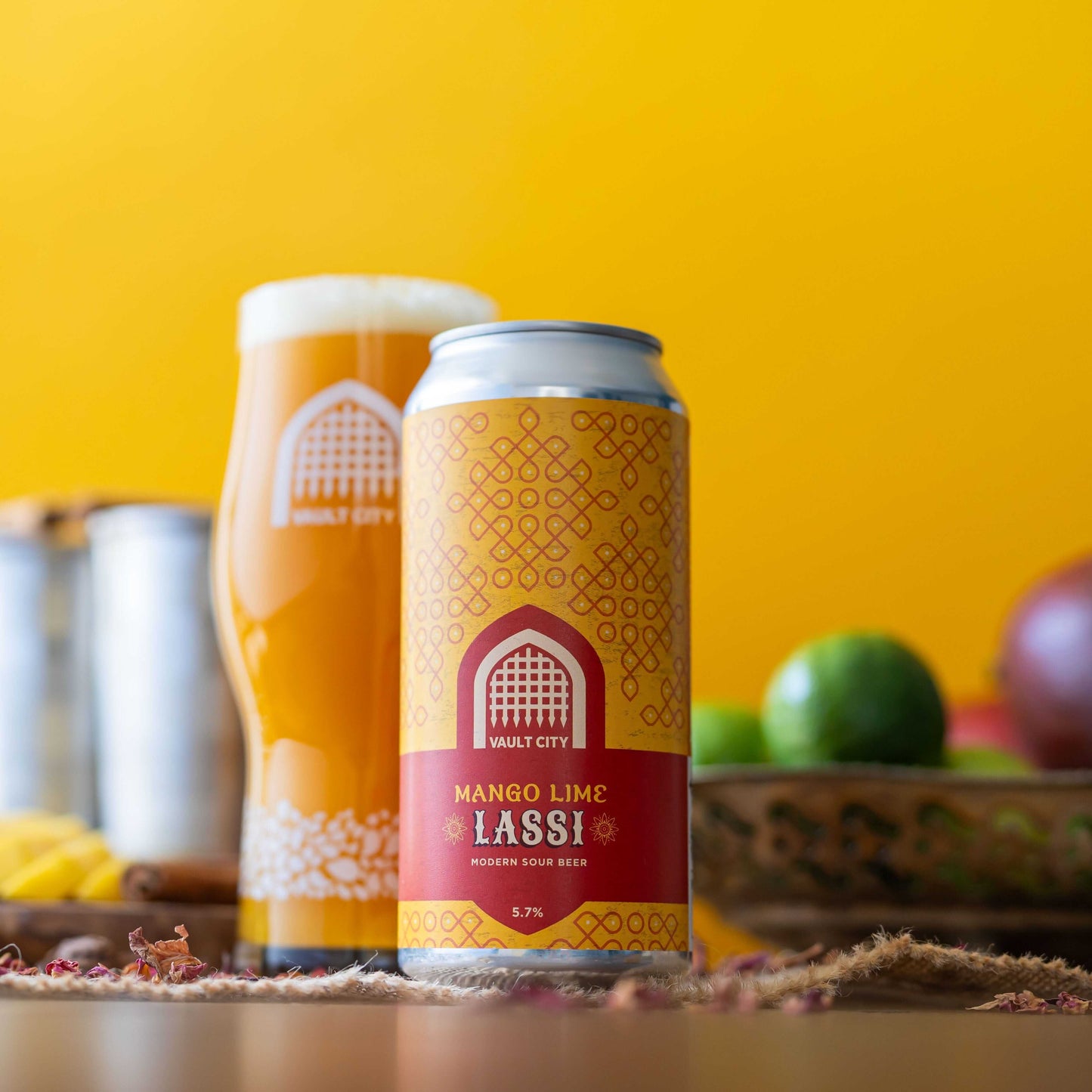 Can of Mango Lime Lassi with fresh mangoes and limes in the background, showcasing juicy mango, vanilla, chai spices, and zesty lime flavors.