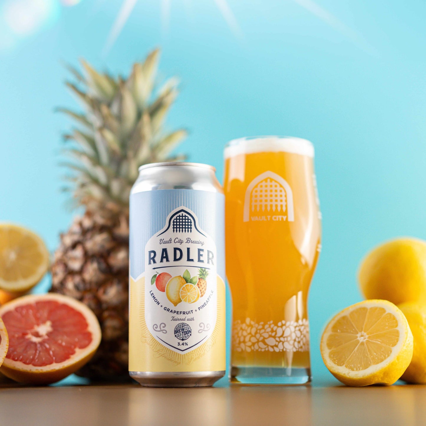 Vault City x Brew Toon Lemon Grapefruit Pineapple Radler can and glass surrounded by fresh fruits in a bright setting