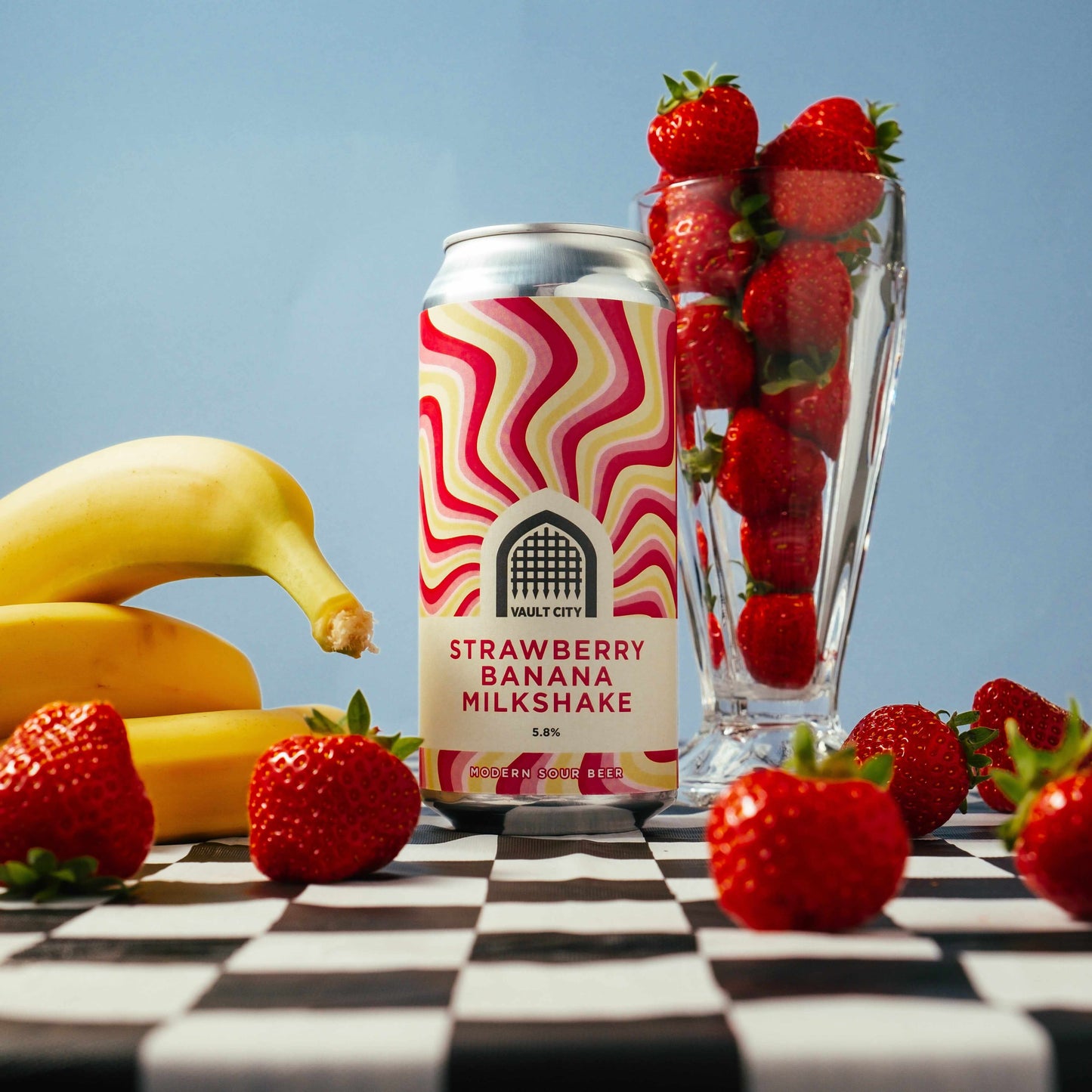 Can of Strawberry Banana Milkshake beer surrounded by fresh strawberries and bananas on a checkered table.