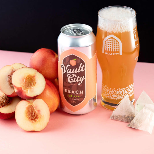 Vault City Peach Ice Tea can and glass with fresh peaches and tea bags on a pink surface.