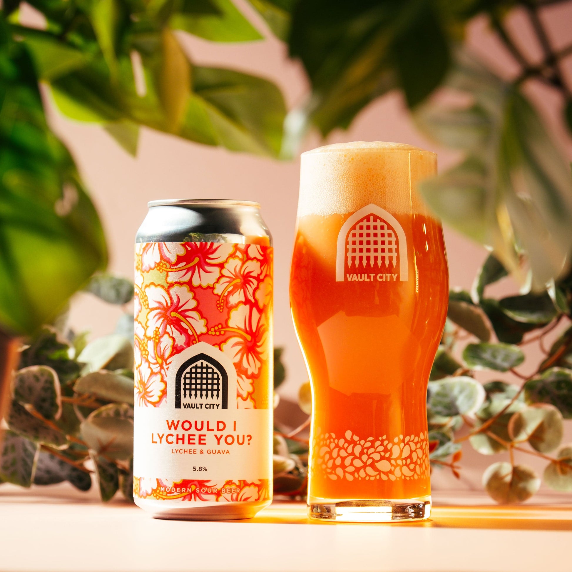 Vault City "Would I Lychee You?" lychee and guava beer with bright pink color in a can and pint glass surrounded by green foliage.