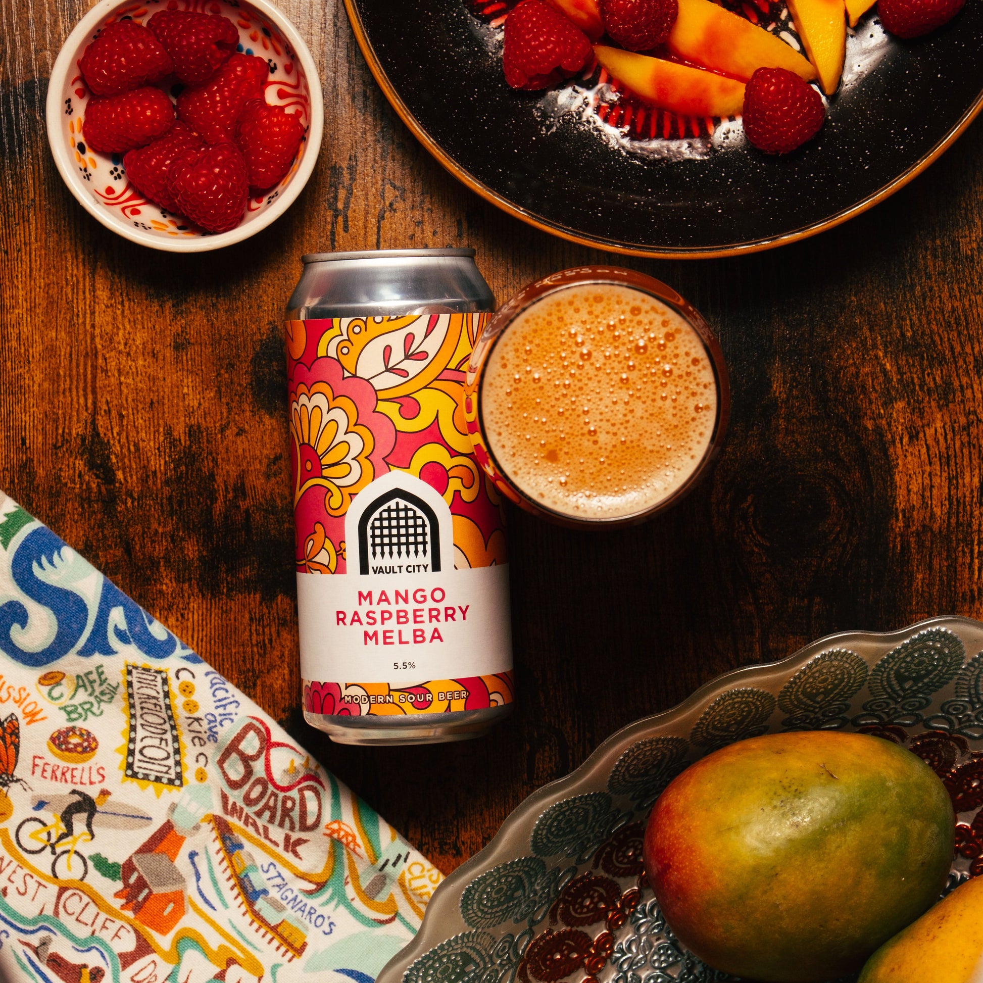 Mango Raspberry Melba sour beer can with fruit, showcasing juicy mango and fresh raspberries, on a wooden table.